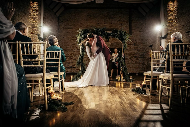 Bride and Groom kissing in front of a wintery floral arch indoors, lit by bright white lights that look like stars at the Great Barn, Aynho.